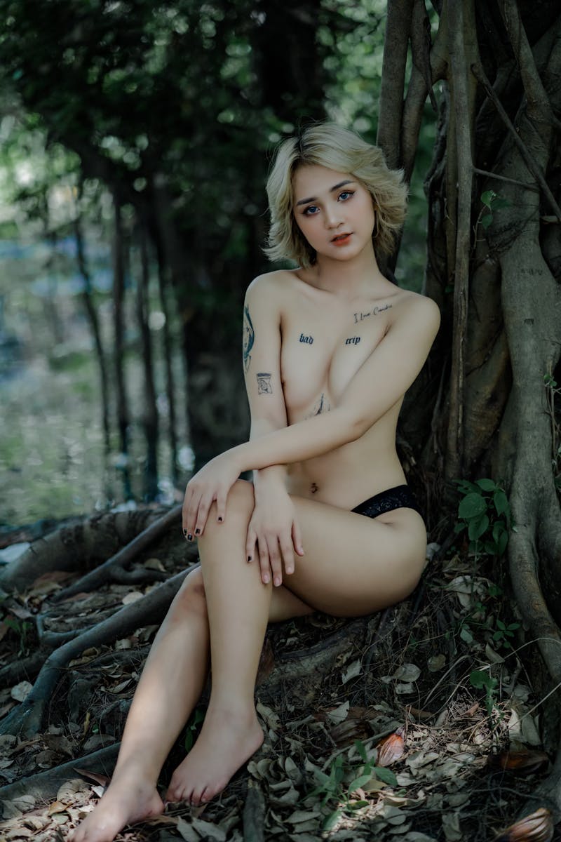 Elegant nude portrait of a tattooed woman sitting by tree roots, exuding natural beauty and artistry.