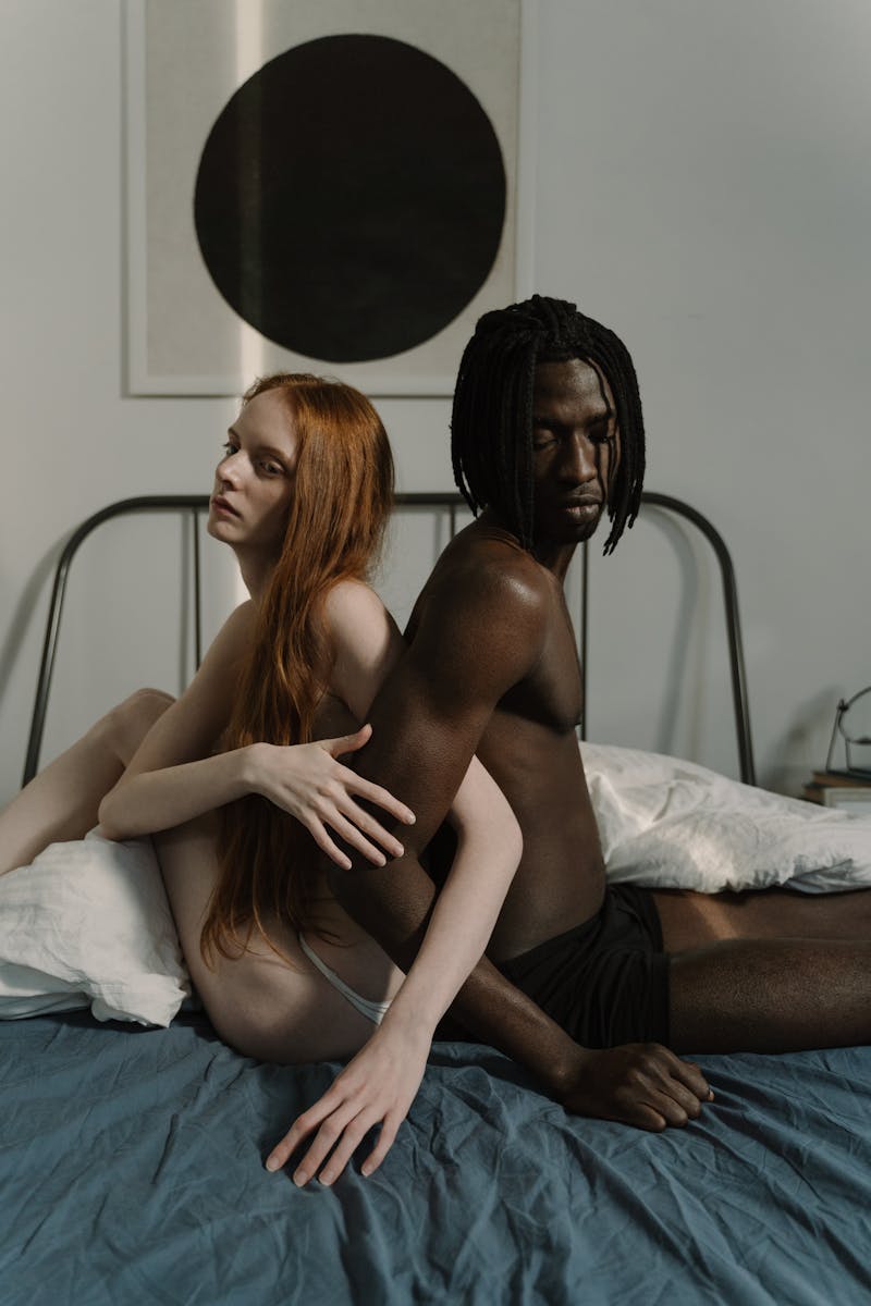 A tender moment captured between a multiracial couple sitting closely in an intimate bedroom setting.